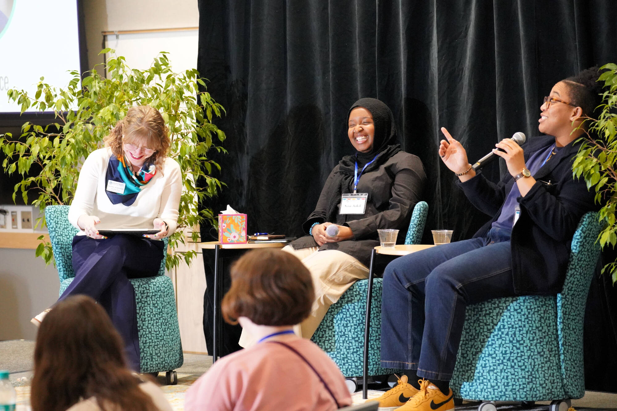 Three speakers on a stage laughing together while one speaks into a microphone.